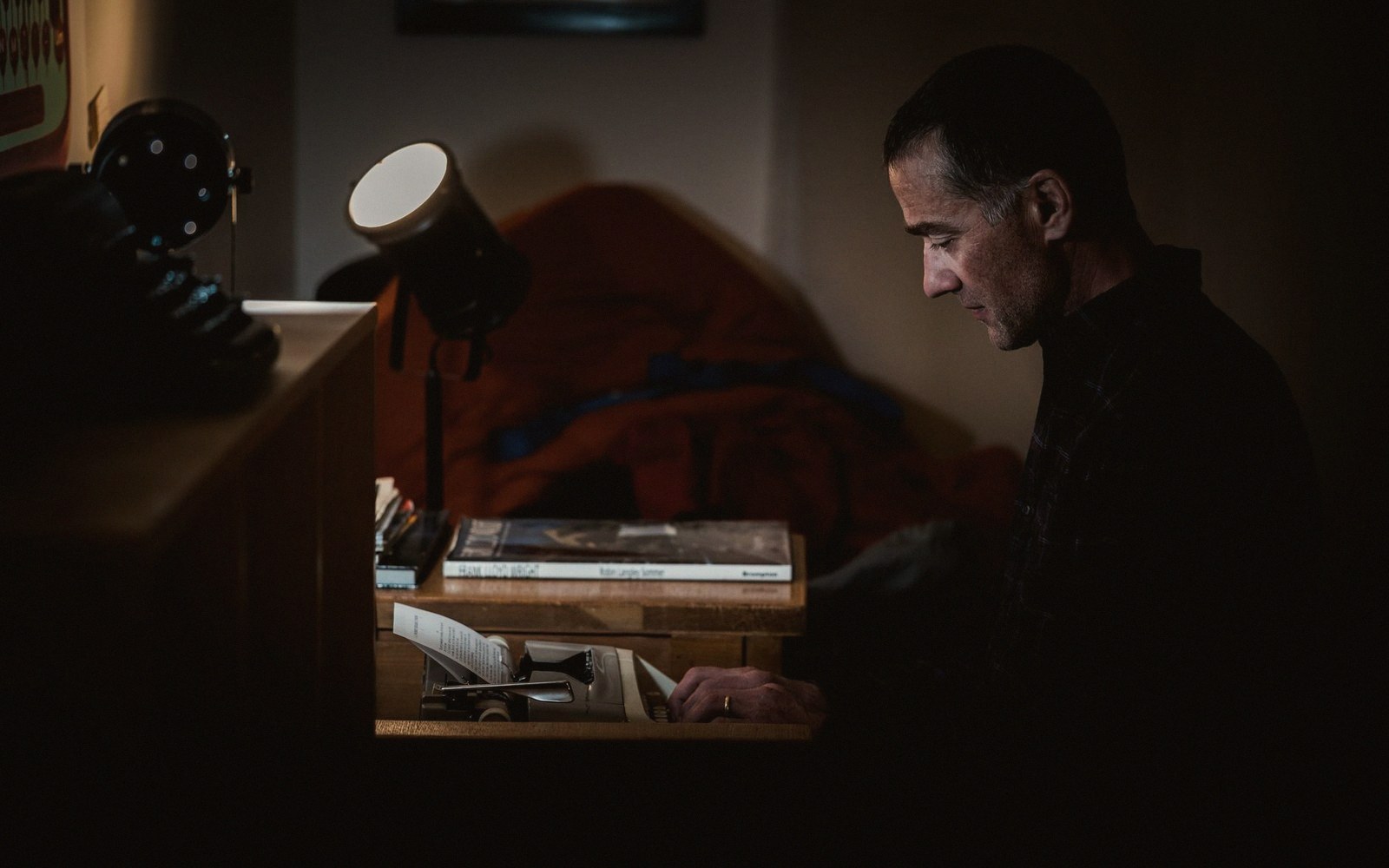A man sits at a desk typing on a typewriter in a dimly lit room