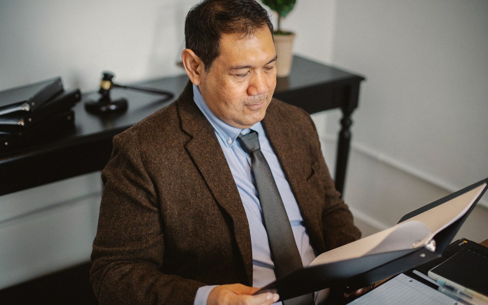 A man in a brown blazer reviews documents in a binder near a gavel
