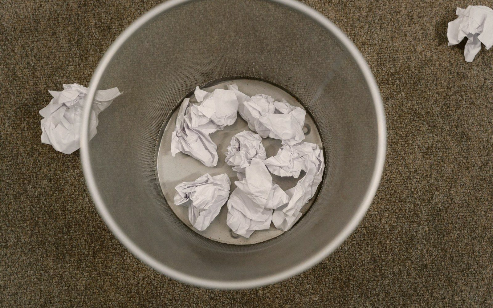 Crumpled balls of paper fill a metal wastebasket, with more scattered on the carpet nearby