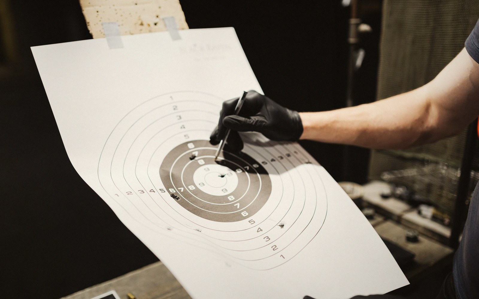 A gloved hand holds a pen near bullet holes on a shooting range target
