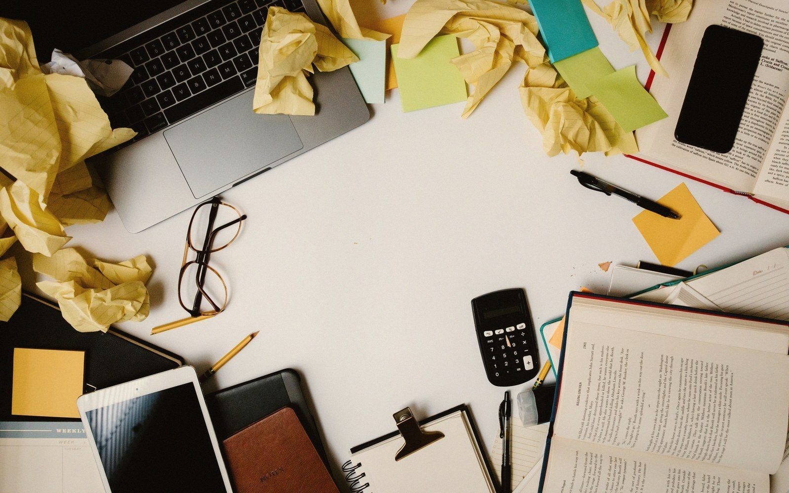 Cluttered desk with laptop, glasses, calculator, notebooks and crumpled paper framing empty workspace