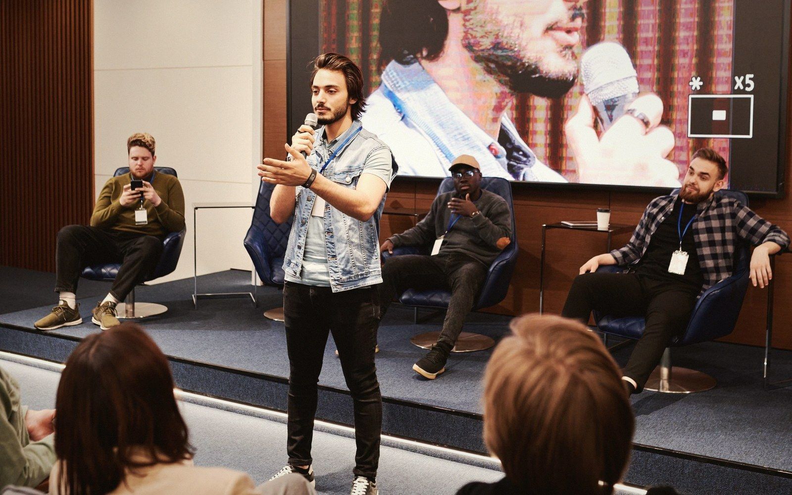 Man speaks into a microphone on stage while panelists sit behind him at a conference