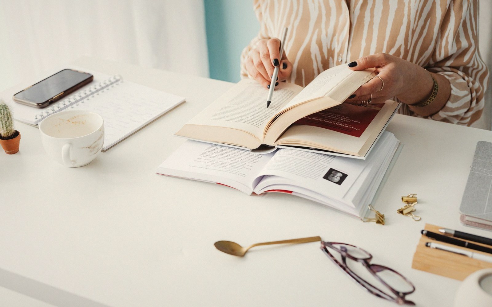 A person annotates an open book with a pen at a desk with coffee and glasses