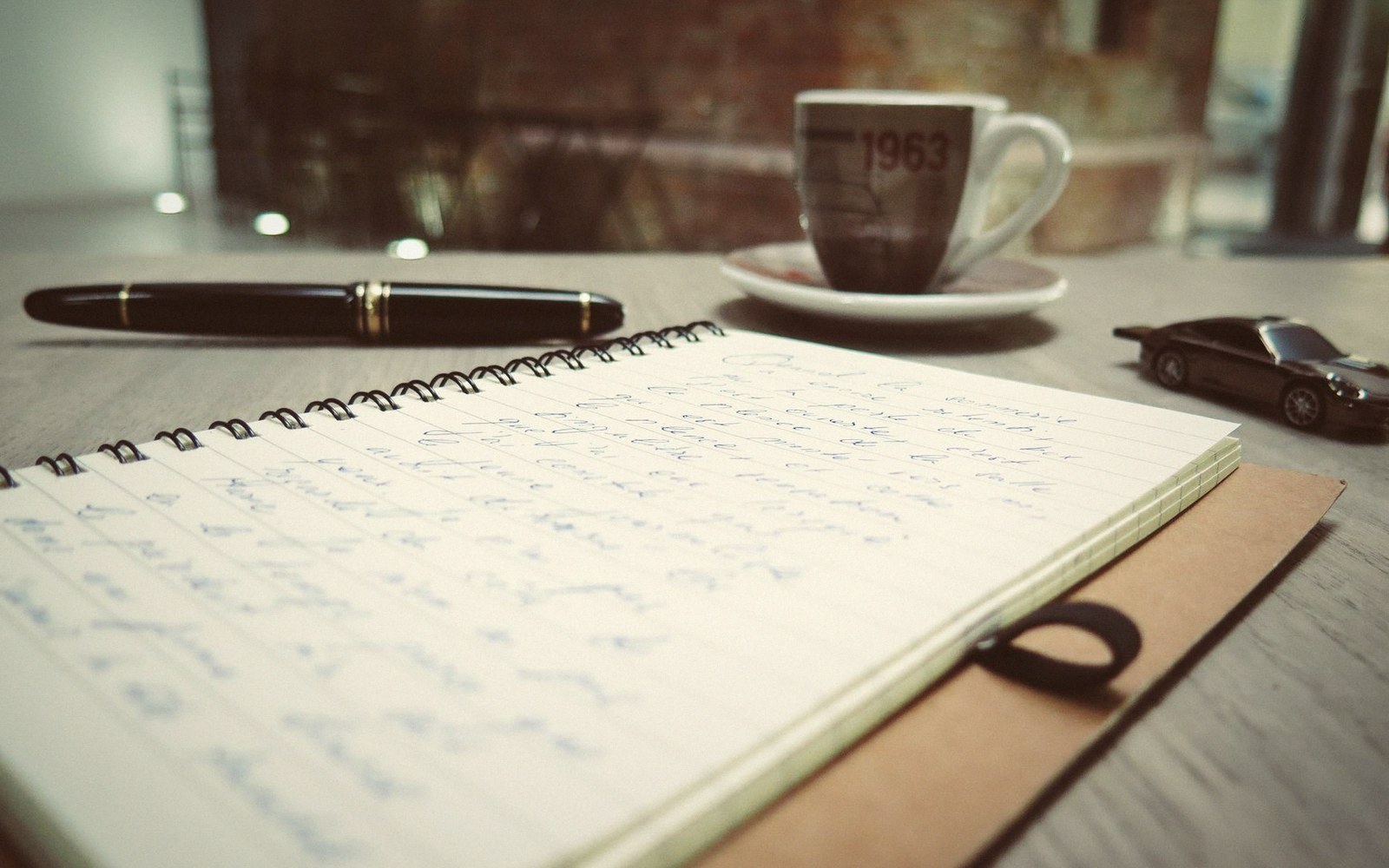 A handwritten notebook, fountain pen, coffee cup and toy car on a wooden table